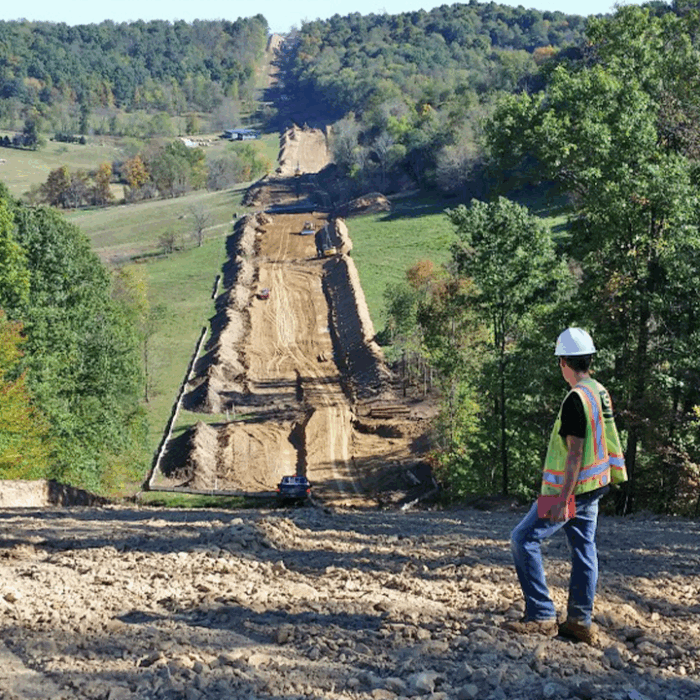 An environmental inspector wearing a safety vest and hard hat observes a large construction site with heavy equipment working along a cleared, graded path through a hilly, forested area.