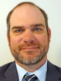 Headshot of a man with short brown hair, a beard, and blue eyes, wearing a suit jacket, dress shirt, and striped tie, smiling slightly against a plain background.