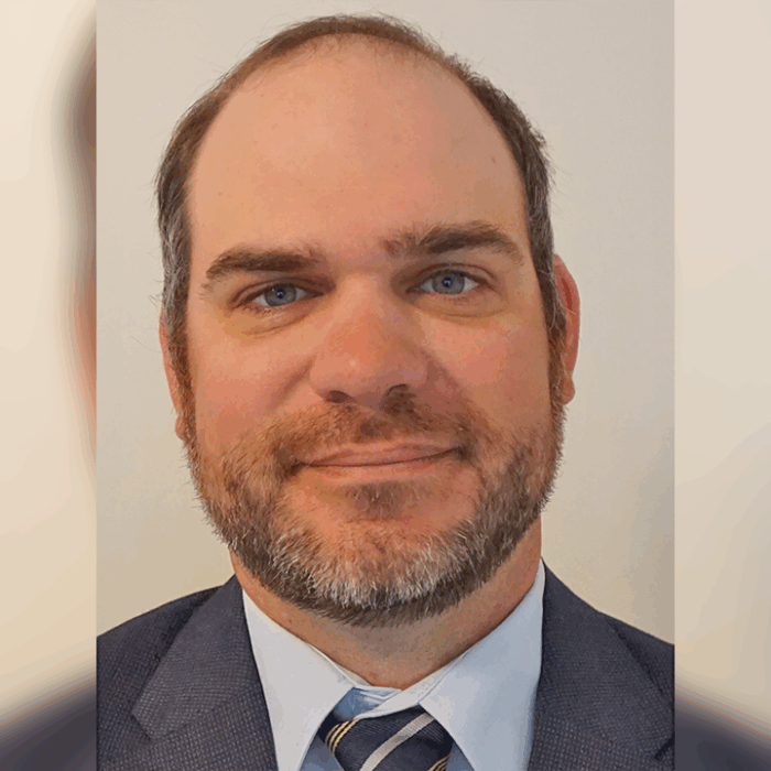 Headshot of a man with short brown hair, a beard, and blue eyes, wearing a suit jacket, dress shirt, and striped tie, smiling slightly against a plain background.
