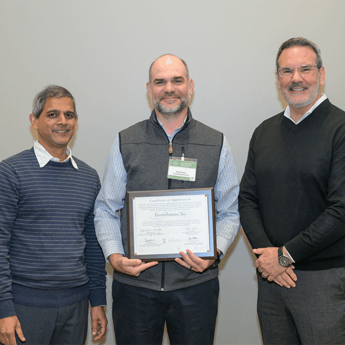 Three professionals stand indoors holding a framed certificate of appreciation, with an EnviroScience representative at center, recognizing the company’s long-standing support and sponsorship at the Railroad Environmental Conference.