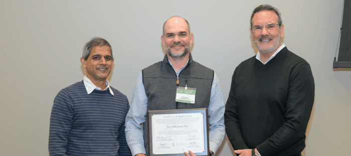 Three professionals stand indoors holding a framed certificate of appreciation, with an EnviroScience representative at center, recognizing the company’s long-standing support and sponsorship at the Railroad Environmental Conference.