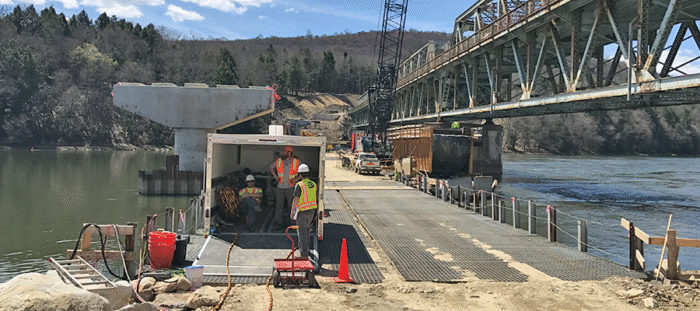 Utility workers conducting maintenance at a hydropower facility along a river beneath an industrial bridge structure.