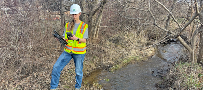 Field scientist in safety vest and hard hat collecting water quality data from a stream using monitoring equipment.