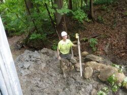 Jeff Niehaus stands in a muddy streambank holding a survey rod, conducting environmental field measurements in a wooded area.