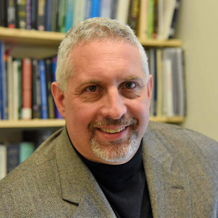 Portrait of Dr. Alan Christian smiling in front of a bookshelf, wearing a blazer and black turtleneck.